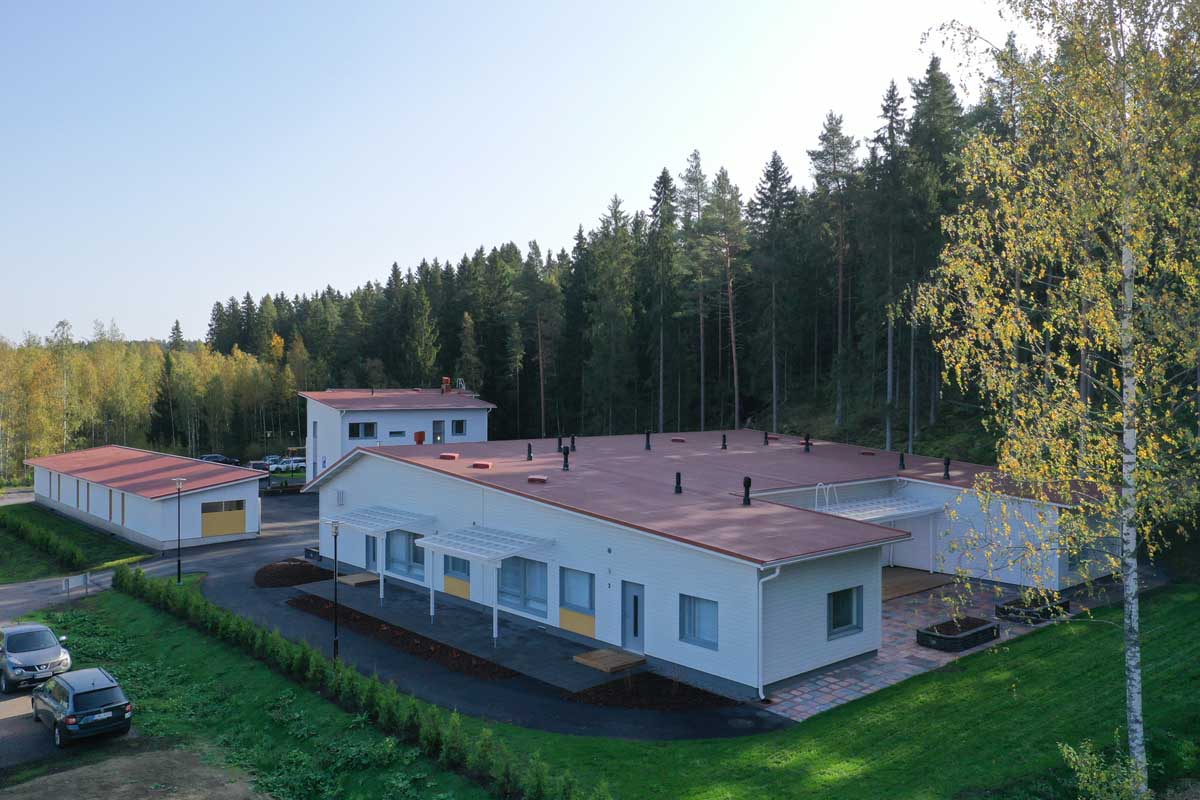 Kuvassa on valkoinen yksikerroksinen perhekuntoutuskeskusrakennus, jossa on punainen katto, vihreä nurmikko ja taustalla metsä.